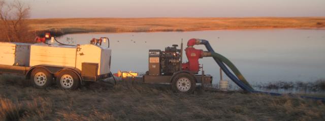 Photo. Daytime. A red-colored, tow-behind, high-volume water pump is positioned near the BR-43 dam’s reservoir. Two large-capacity, black-colored, flexible pipes transfer water from the reservoir to the pump and from the pump to a designated flow channel (not pictured). Blue skies, sunny, mix of dried and green grasses in the foreground. 