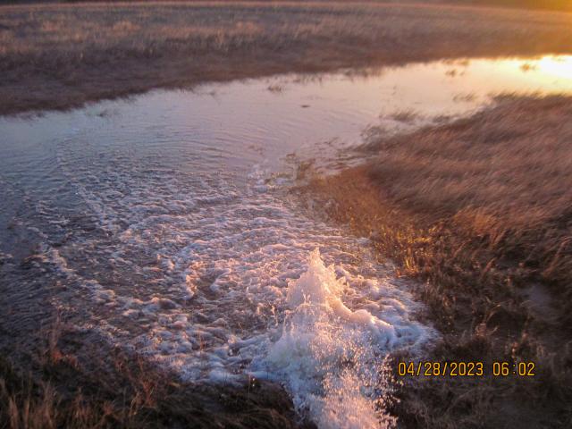 Photo. Daytime. Water diverted from the reservoir to a designated flow channel. Sunshine on a mix of dried and green grasses on all sides of the flowing water.