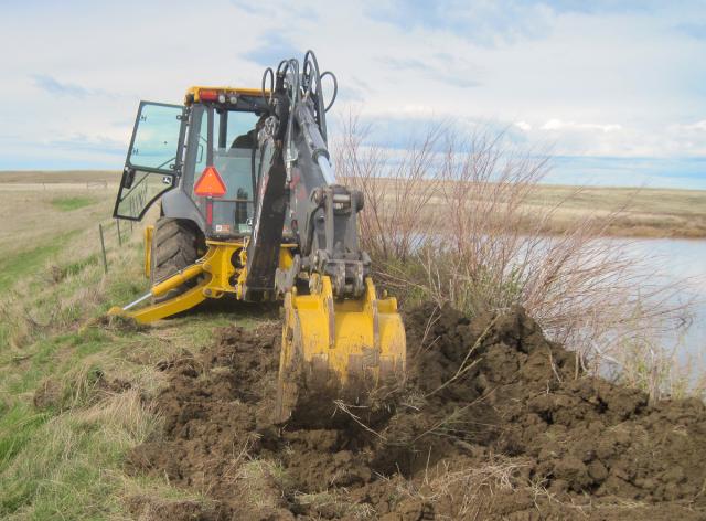 Photo. Daytime; blue, partly cloudy skies. A yellow-colored construction excavator creates a trench in the ground to temporarily repair the point of seepage and piping. Mix of dried and green grasses on the ground.