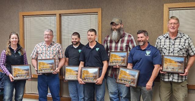 Photo. Indoor office setting. Seven people stand along a wall with two windows, blinds closed, holding framed, picturesque award certificates.