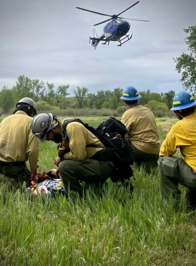 Four people in yellow shirts and blue hard hats crouch in the grass as a blue helicopter lands nearby.