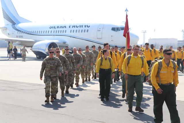 A line of people dressed in camouflage and a line of people in yellow shirts and green pants march away from an airplane.