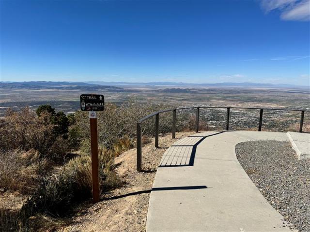 The photo shows a sidewalk with a fence on the left overlooking an expansive landscape with a clear blue sky.