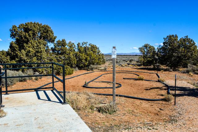 The photo shows the corner of a cement platform with metal railing, which is overlooking a dirt track for radio-controlled cars. The track loop is surrounded by juniper trees and sagebrush under a deep blue sky. 