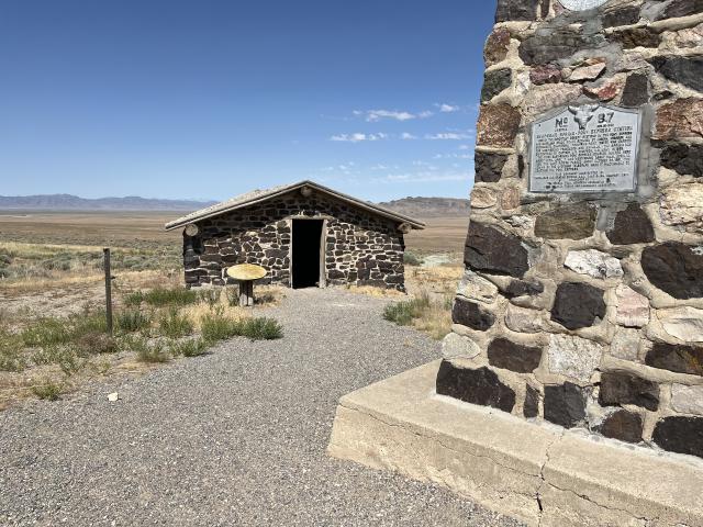 The photo shows a rock monument with an informational metal plaque, next to that is a fine gravel pathway leading into a stone building, which was a Pony Express station. There’s a metal fence post and old round table in front of the building   