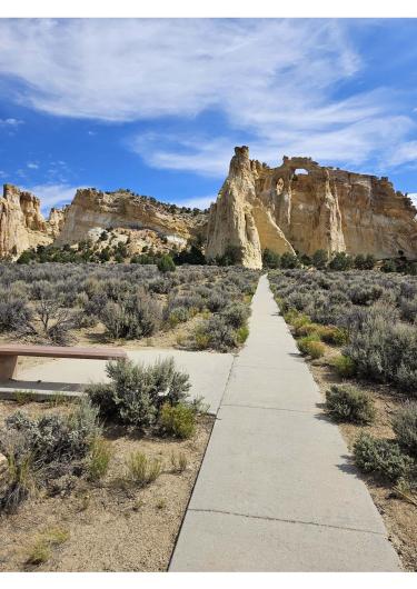 This photo shows a sidewalk with a pull-out on the left with a bench, leading to a sandstone arch. It goes through a dry, low shrub landscape and sits under a wispy-cloud-filled blue sky. 