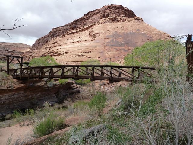 The photo shows a dark brown metal bridge spanning a small canyon with a large sandstone rock formation in the background. 