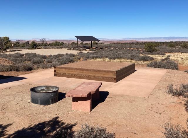 The photo shows an elevated wood tent pad surrounded by a concrete path in the center of the frame, with a flagstone bench and fire ring in front of it. A gravel parking lot and shaded picnic table are in the background, under a blue sky and surrounded by low shrub-grassland.