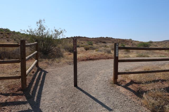 This photo shows the beginning of a compact gravel trail. A vertical pole bisects the trail, which bends to the right. A 3-pole fence is on either side of the trail perpendicular to the trail and lined up with the single vertical pole.  