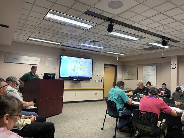 A group of people sit in a conference room while a Caucasian man speaks from behind a podium next to a projected presentation. 