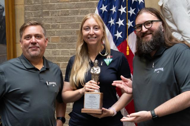 Two Caucasian men and a Caucasian woman pose smiling with a silver trophy shaped like Smokey Bear.