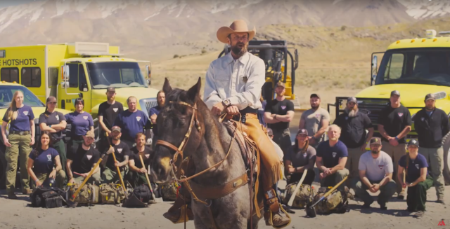 A Caucasian man in cowboy clothes riding a horse speaks in front of posed modern wildland firefighters and engines