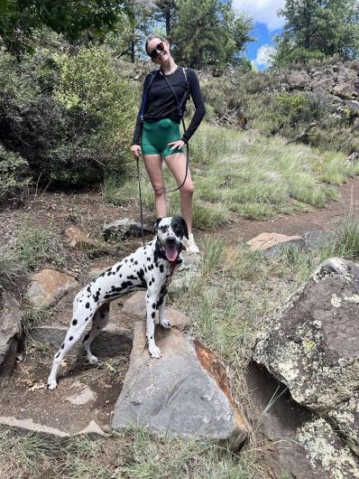 The photo shows a young woman and a dalmatian dog standing on a dirt trail heading up a hill. There are lichen stained basalt rocks, grasses, shrubs, and small evergreen trees next to the trail.