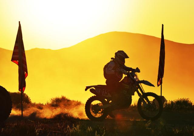 Silhouette of a rider on a dirtbike creating a dust cloud between two large posted flags in an orange horizon