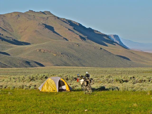 A lone tent and dirt bike sit in a grassy area backdropped by a low hillside