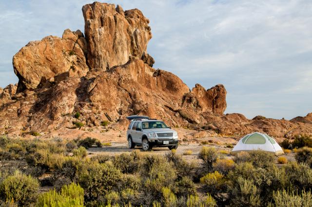 A lone vehicle and tent surrounded by sagebrush and a dramatic rock formation.