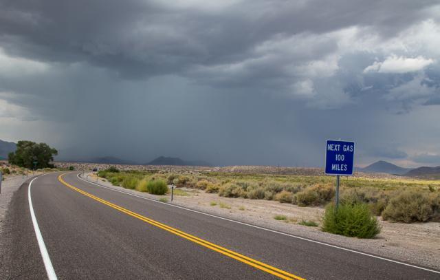 An empty highway curves past a sign that reads Next Gas 100 miles backdropped by a dense grey storm cloud.