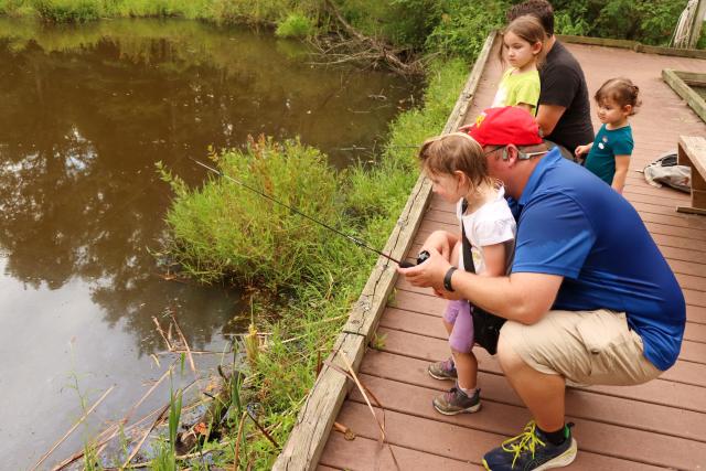 A man crouches on a dock behind his young daughter, helping her hold a fishing rod on a dock along a pond. 