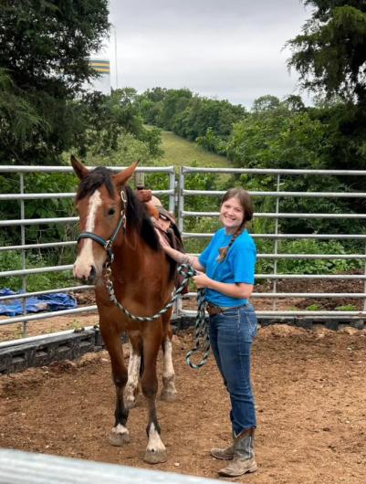 A woman smiles at the camera as she stands next to her brown horse with white facial markings inside of an outdoor pen