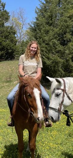 A woman rides on a brown horse, as a white horse's head is ivisible coming in from the right side of the phot, with green pine trees and blue sky visible in the background.