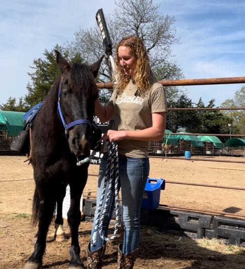 A woman smiles as she stands next to a black horse. 