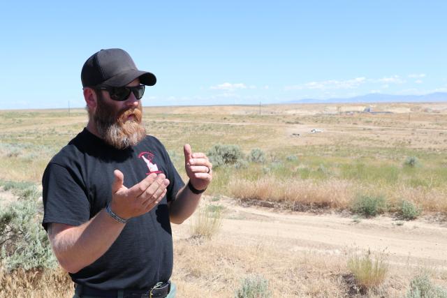 Caucasian man in dark ballcap and tshirt talks and gestures outdoors next to a flat grassy plain and dirt road