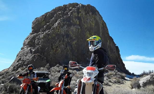 Three dirt bike riders in full gear pose in front of a large rock landmark.