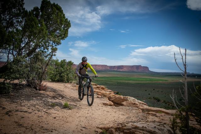 A mountain bike rides on a single track path in front of sweeping green mesas.