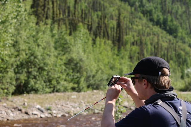 Man assessing a river in front of green trees and a soily riverbank.