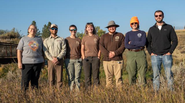 Seven people stand in a row smiling at the camera. They are outside in a grassy field facing the sun.