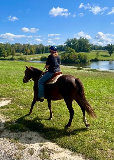 A woman rides a brown horse away from the camera on a green grassy field, a small pond and trees in the background. 