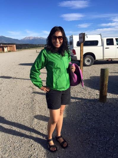 Wearing a green jacket and sunglasses, Vanessa Lacayo stands in a gravel parking area under a bright blue sky. A snow-covered mountain can be seen, in the distance, over her right shoulder.