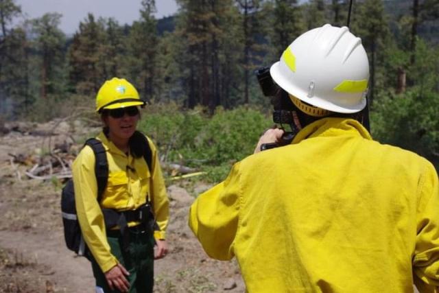 Vanessa Lacayo responds to a question while being interviewed near the site of a wildland fire. She is dressed in yellow and green BLM fire gear, equipped with a safety helmet, sunglasses, and a backpack.