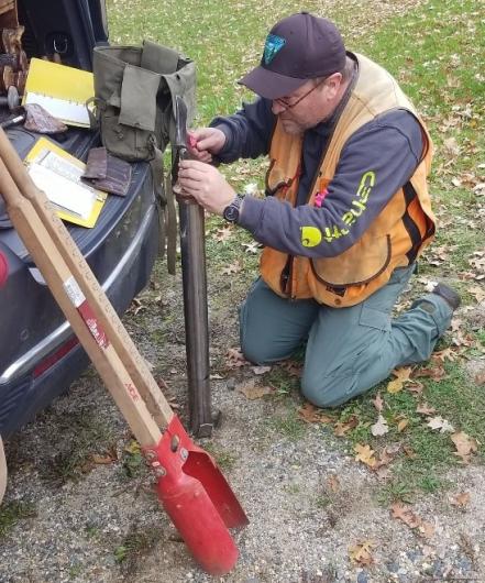 A survey team member kneels as he works on tools leaning against a vehicle. 