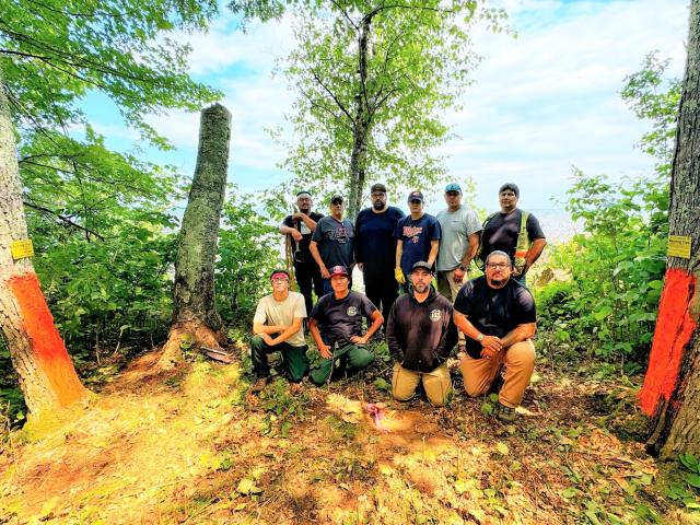 A group photo of 10 men, 4 kneeling on the ground, with 6 standing, trees are on either side. 