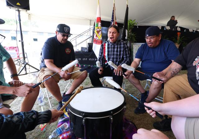 Underneath a large tent, six men sit around a large drum, all holding mallets, as one sings. 