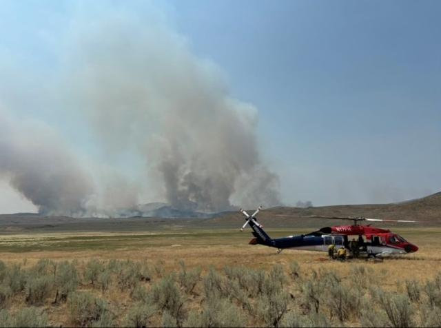 A helicopter landed in a field with light gray smoke rising from hills in the distance