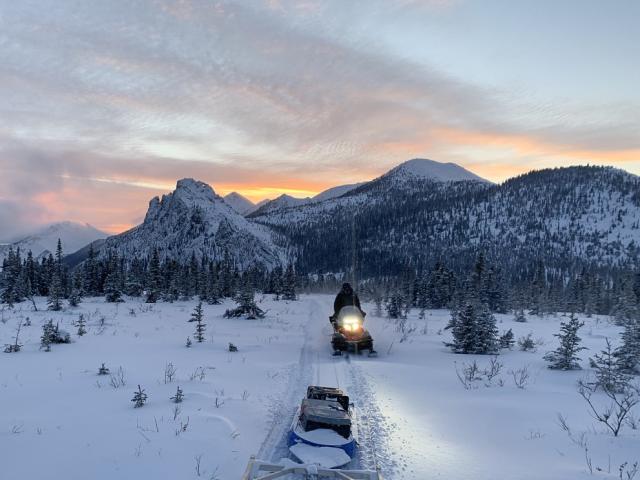 Looking behind a snowmobile over the sled with gear at another rider on a snowmobile silhouetted as the sun sets. 