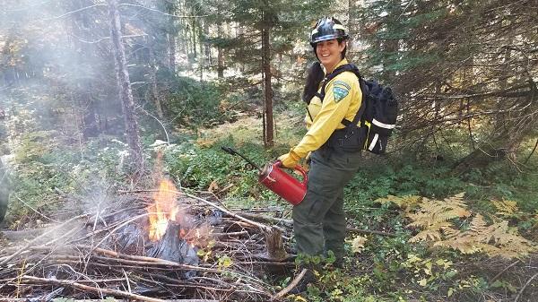 smiling firefighter in traditional yellow shirt holding a drip torch near a burn pile