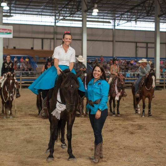 A person in a blue shirt and blue skirt sits on top of a dark brown horse and a person in a blue shirt and black pants stands next to them.  