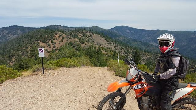 A dirt bike rider in the foreground looks over hills of chaparral and a lake