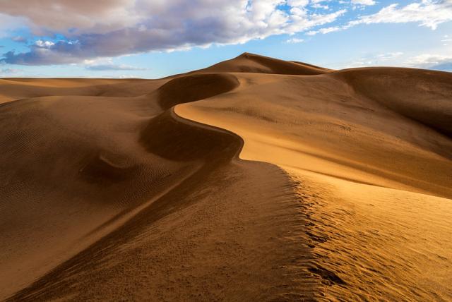 The ridge of a sand dune winds upward toward a cloudy sky.