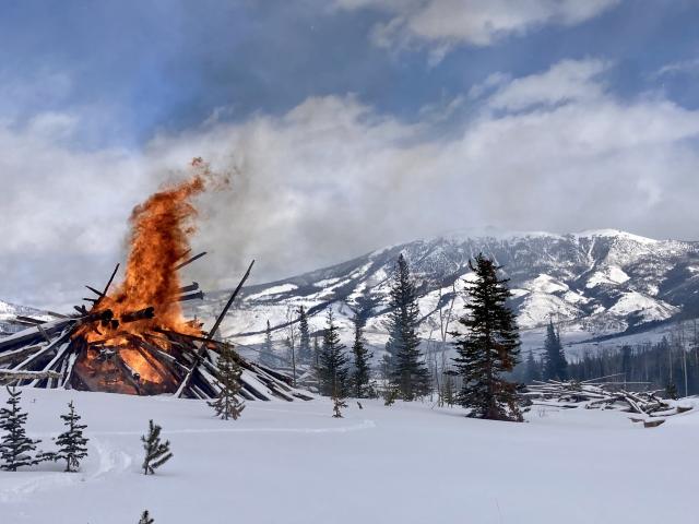 Winter scene with snow on ground and mountain backdrop with large pile of trees and debris aflame