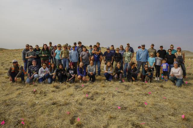 A large group of students and adults pose for a group photo in an open, grassy area with small pink flags in the ground in front of them.