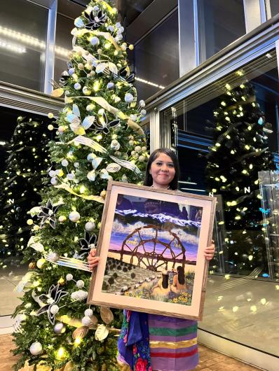 A girl with long black hair and colorful dress smiles holding a framed colorful piece of artwork