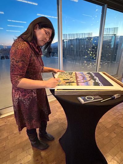 A woman with long dark hair stands signing an art poster.