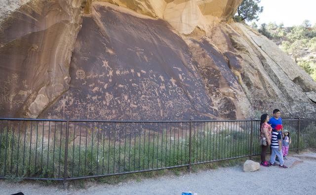 Three people, including a child stand in front of a petroglyph panel and a metal fence.