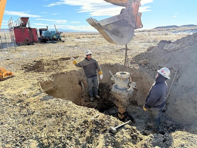 Two men stand nearly shoulder deep in a large hole in the ground watching a heavy load dangles on a chain from a loader bucket. Large pile of dirt and other equipment in the background.