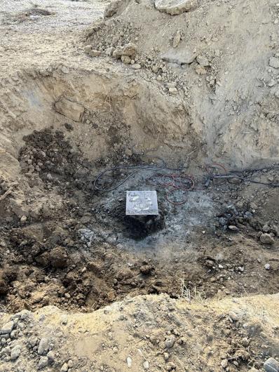 A square piece of metal with writing in the bottom of a large hole in the dirt. Various cords are coiled nearby. 