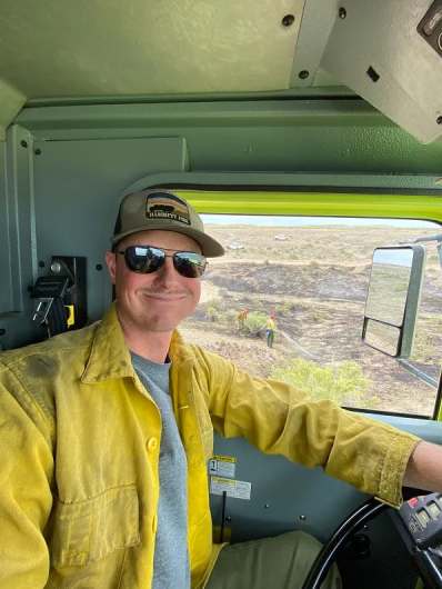 Caucasian man in sunglasses and a yellow shirt smiles sitting in the cab of a truck.